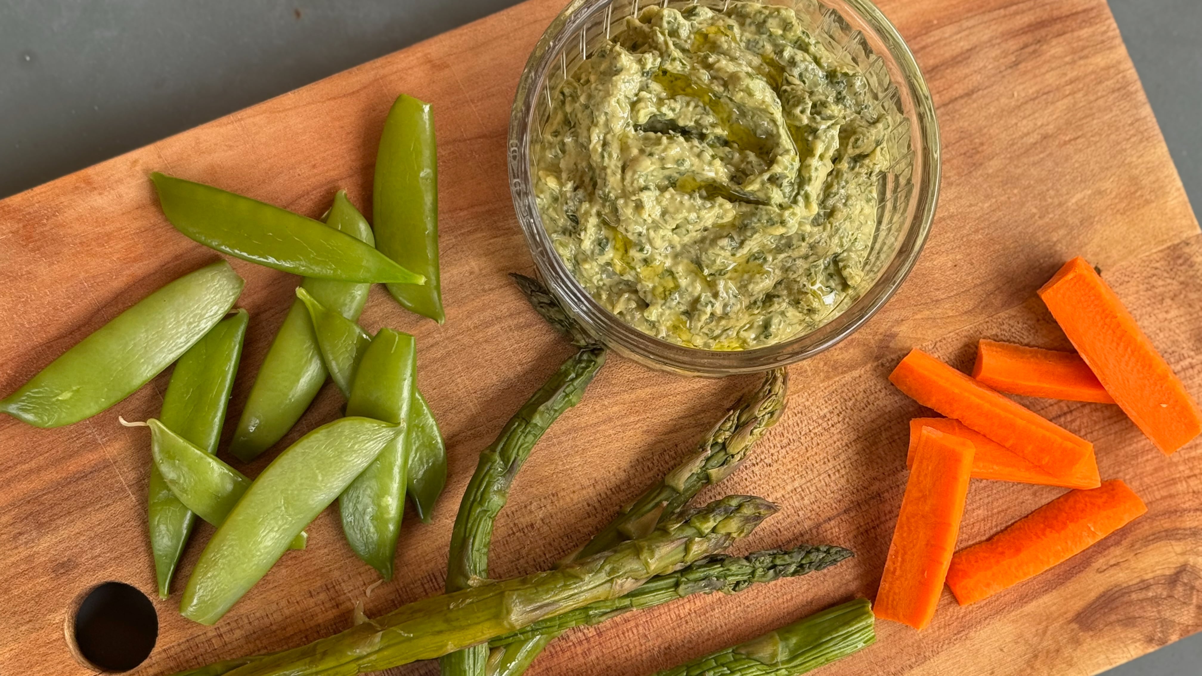 a bowl of green tahini on top of a cutting board alongside carrots, asparagus and snap peas